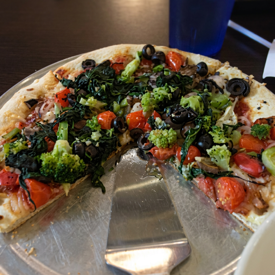 Vegetable pizza with broccoli, tomatoes, olives, and melted cheese on a metal tray.